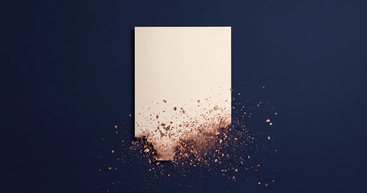 A single sheet of cream paper on a deep navy field, seen from above, intact along its upper edge while its lower portion breaks apart into scattered copper fragments and dust
