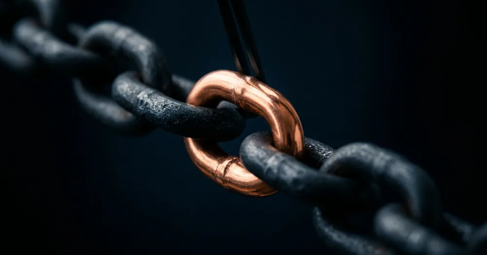 A polished copper chain link held by tongs, being fitted into a long chain of dark steel links
